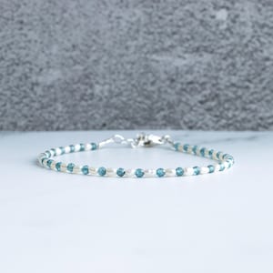 Könnte beinhalten: Zartes Armband mit kleinen, runden, hellblauen Perlen und weißen Perlen. Das Armband ist auf einem dünnen Band aufgereiht und hat einen silbernen Verschluss. Der Schmuck wird auf einer weißen Oberfläche präsentiert.