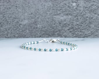 May include: A delicate bracelet with small, round, light blue beads and white beads. The bracelet is strung on a thin cord and has a silver clasp. The jewellery is displayed on a white surface.