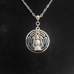 Könnte beinhalten: Eine silberne Halskette mit einem Anhänger, der eine stilisierte Laterne in einem kreisförmigen Labyrinth zeigt.