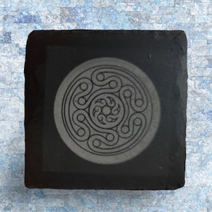 May include: Square black slate coaster with a Celtic knot design etched in the center. The coaster has a natural, rough edge and a light gray circular design. The background is a blue and white textured surface.