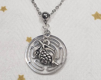 Puede incluir: Un collar de plata con un colgante circular que presenta un diseño de granada estilizado. El colgante está rodeado por un patrón de círculos concéntricos.