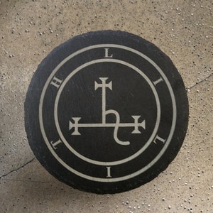 Puede incluir: Posavasos redondo de pizarra negra con un diseño gráfico blanco. El diseño presenta un borde circular con las letras H, L, I y T, y un símbolo central con elementos en forma de cruz y una línea curva. El posavasos está sobre una superficie texturizada.