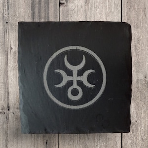 May include: Square black slate coaster with a light gray etched design. The design is a circular emblem with a central cross-like shape and crescent moon shapes. The coaster sits on a wooden surface.