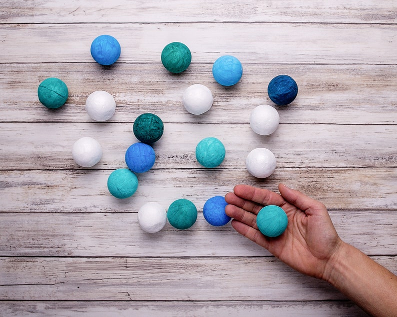 The balls of the DIY Montessori Gobbi mobile kit for newborn visual development, on a rustic wooden surface, with a hand holding a ball for scale