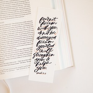 May include: A white bookmark with black calligraphy that reads "Fear not, for I am with you; do not be dismayed, for I am your God. I will strengthen you and help you." The text is in a modern calligraphy style with a few orange accents. The bookmark also includes the verse "Joshua 1:9-10" in smaller font.
