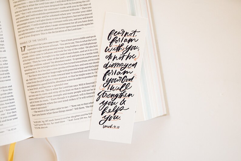 May include: A white bookmark with black calligraphy that reads "Fear not, for I am with you; do not be dismayed, for I am your God. I will strengthen you and help you." The text is in a modern calligraphy style with a few orange accents. The bookmark also includes the verse "Joshua 1:9-10" in smaller font.