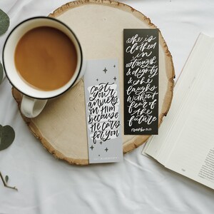 May include: Two black and white bookmarks with inspirational quotes. The quote on the left bookmark reads "Cast your anxiety on Him because He cares for you." The quote on the right bookmark reads "She is clothed in strength and dignity and she laughs without fear of the future."  The bookmarks are on a wooden surface with a cup of coffee and a book.