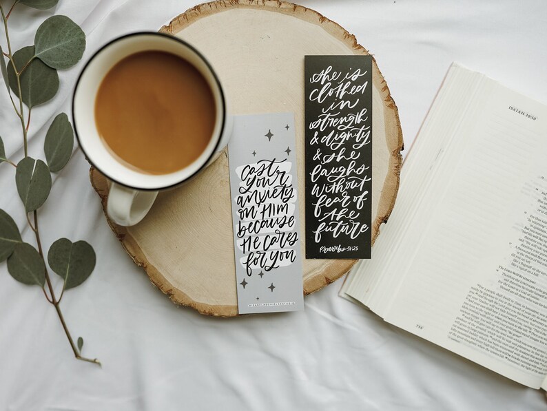 May include: Two black and white bookmarks with inspirational quotes. The quote on the left bookmark reads "Cast your anxiety on Him because He cares for you." The quote on the right bookmark reads "She is clothed in strength and dignity and she laughs without fear of the future."  The bookmarks are on a wooden surface with a cup of coffee and a book.