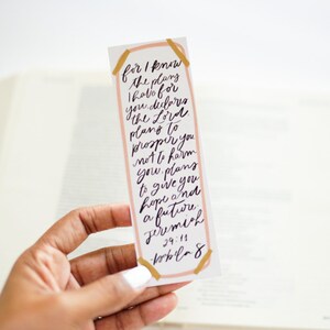 May include: A pink and gold bookmark with a handwritten quote from Jeremiah 29:11, "For I know the plans I hold for you, declares the Lord, plans to prosper you and not to harm you, plans to give you hope and a future."