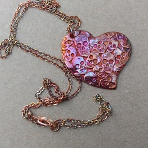 May include: A heart-shaped pendant necklace with a copper finish. The pendant is covered in a pattern of tiny skulls. The necklace is made of a multi-colored chain.