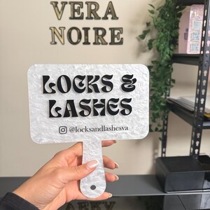 May include: A white and black sign with the text "LOCKS & LASHES" and the Instagram handle @locksandlashesva. The sign is held by a person. The words "VERA NOIRE" are on the wall in the background.