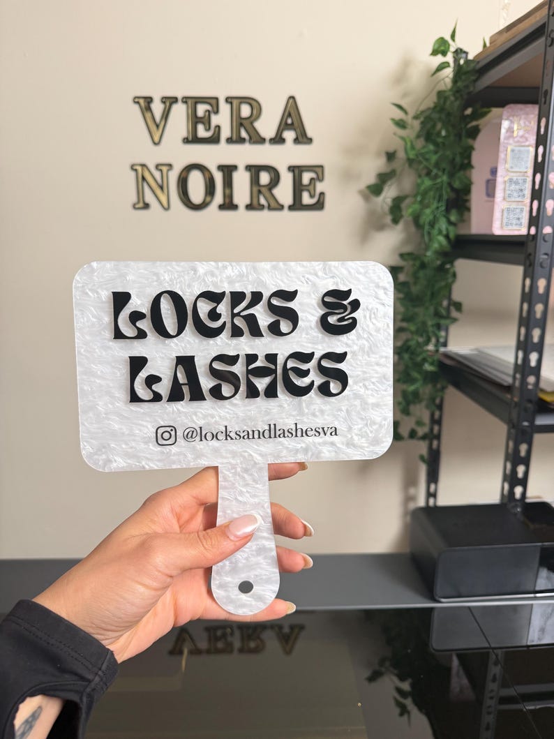 May include: A white and black sign with the text "LOCKS & LASHES" and the Instagram handle @locksandlashesva. The sign is held by a person. The words "VERA NOIRE" are on the wall in the background.