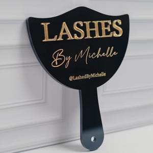 May include: Black sign with gold lettering that reads "LASHES By Michelle" and the Instagram handle "@LashedByMichelle". The sign has a handle and a hole at the bottom.