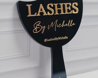 May include: Black sign with gold lettering that reads "LASHES By Michelle" and the Instagram handle "@LashedByMichelle". The sign has a handle and a hole at the bottom.