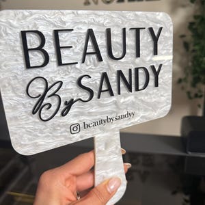 May include: A white and gray marbled acrylic sign with the words "BEAUTY By SANDY" in black lettering. The Instagram handle @beautybysandy is printed at the bottom. The sign is held by a hand.
