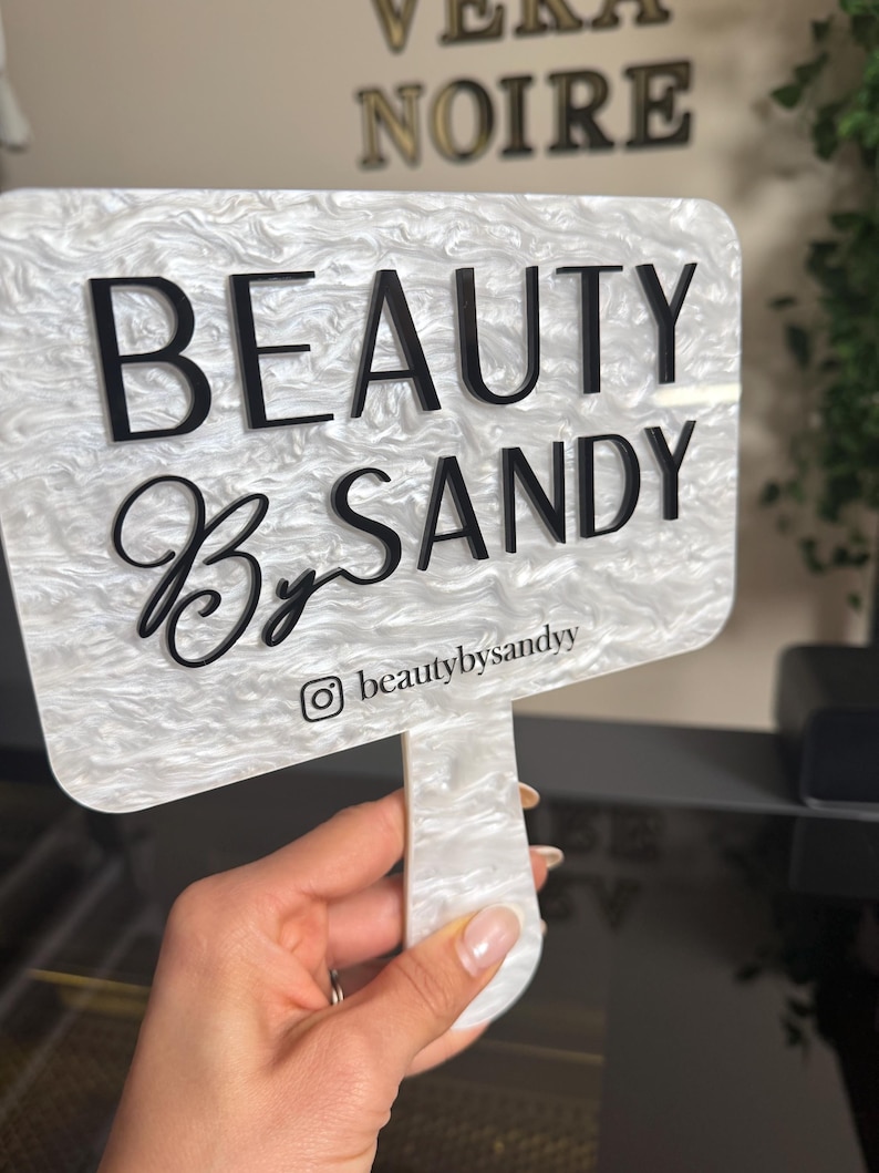 May include: A white and gray marbled acrylic sign with the words "BEAUTY By SANDY" in black lettering. The Instagram handle @beautybysandy is printed at the bottom. The sign is held by a hand.