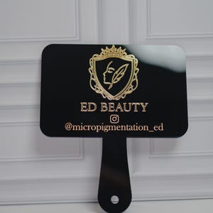 May include: A black sign with a handle, featuring the gold text "ED BEAUTY" and the Instagram handle "@micropigmentation_ed." The sign has a gold emblem of a woman's profile and a feather within a crown.