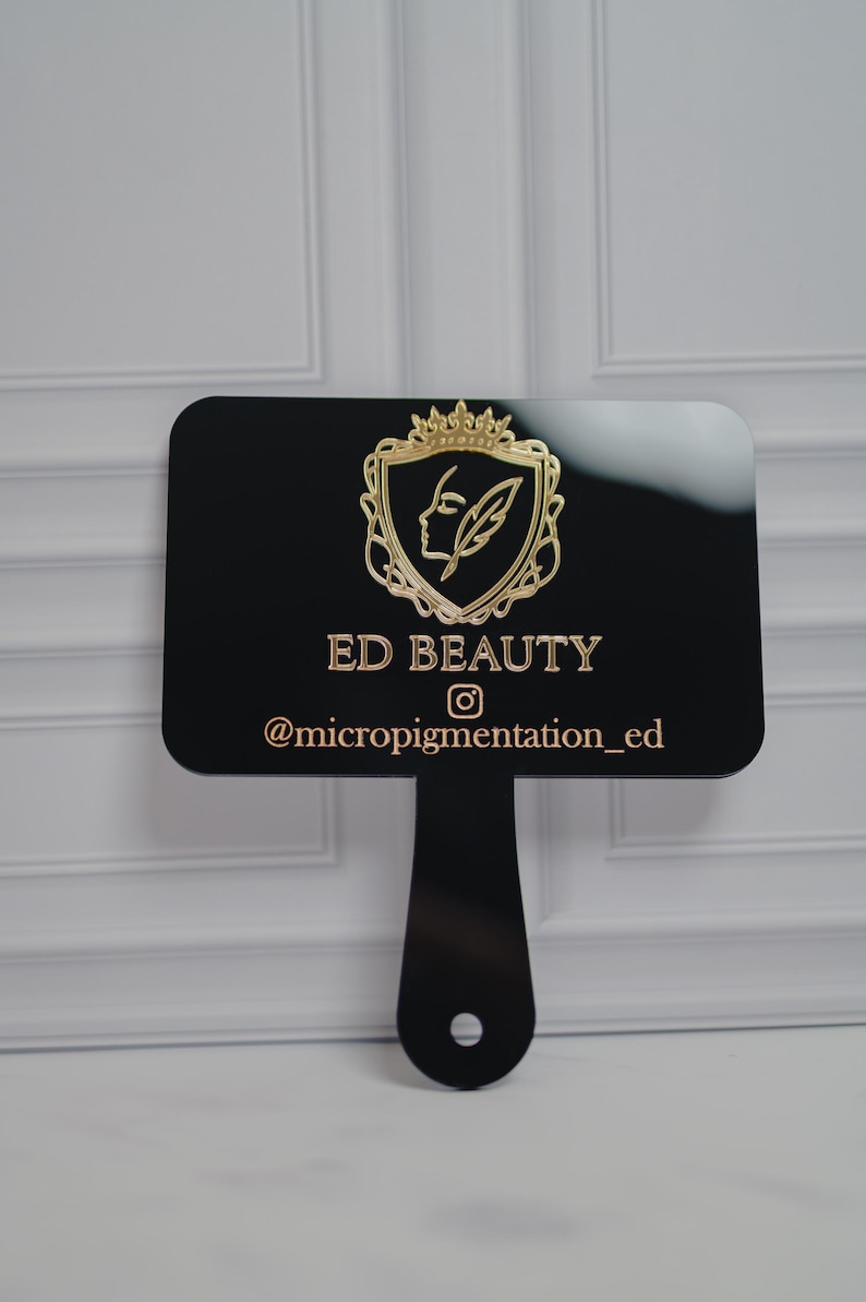 May include: A black sign with a handle, featuring the gold text "ED BEAUTY" and the Instagram handle "@micropigmentation_ed." The sign has a gold emblem of a woman's profile and a feather within a crown.