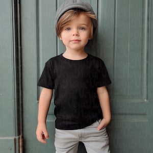 May include: A young child wearing a black t-shirt, gray pants, and a gray newsboy cap. The t-shirt has a repeating pattern of the words "Vera Noire" across the fabric. The child is standing in front of a green door.
