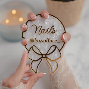 Puede incluir: Un letrero acrílico blanco y dorado con la palabra "Nails" y el nombre de usuario de Instagram "kasallure_". El letrero tiene un diseño de lazo y lo sostiene una persona.