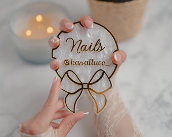 May include: A white and gold acrylic nail art display with the word "Nails" and the Instagram handle "kasallure_". The display has a bow design and is held by a person with manicured nails.