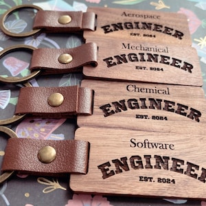 May include: Four wooden keychains with leather straps. Each keychain has a different engineering field title: Aerospace Engineer, Mechanical Engineer, Chemical Engineer, and Software Engineer. Each title is in a curved font with the year 2024 below.