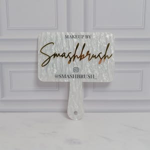 May include: A white, rectangular makeup mirror with a handle. The mirror features the text "MAKEUP BY Smashbrush" in a gold script font, along with the Instagram handle "@SMASHBRUSH_". The mirror has a pearlescent, textured surface.