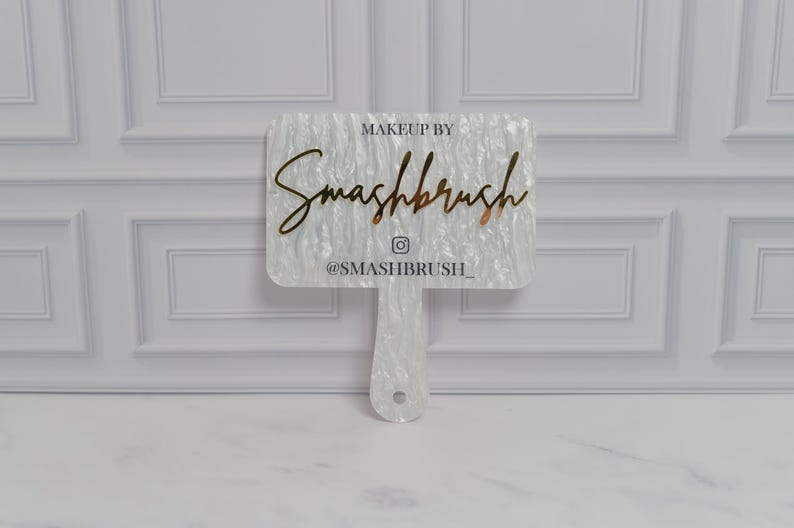 May include: A white, rectangular makeup mirror with a handle. The mirror features the text "MAKEUP BY Smashbrush" in a gold script font, along with the Instagram handle "@SMASHBRUSH_". The mirror has a pearlescent, textured surface.