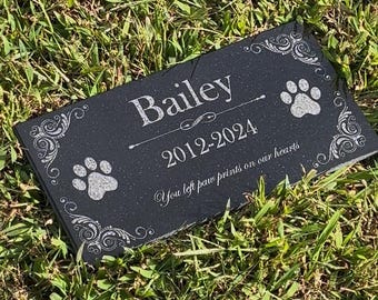 May include: A black rectangular pet memorial stone with the name "Bailey", dates 2012-2024, paw prints, and decorative scrollwork. The inscription reads "You left paw prints on our hearts".