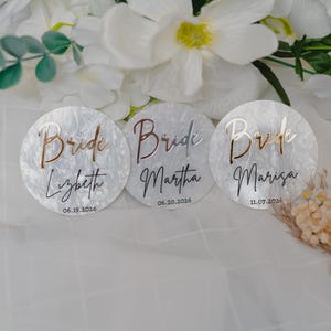 May include: Three circular white marbled acrylic discs featuring the word Bride in metallic script, followed by a name and a date. The first disc reads Lizbeth 06.19.2026, the second Martha 06.20.2026, and the third Marisa 11.07.2026 in black text.