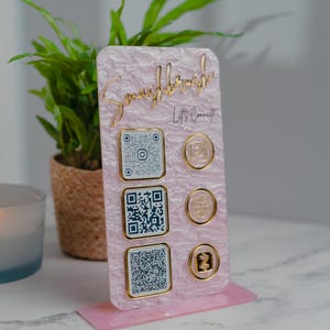 May include: A pink acrylic social media display stand with gold accents. The stand features QR codes and social media icons. The text "Smashbrush" and "Let's Connect" are displayed in gold lettering. A small plant and candle are in the background.