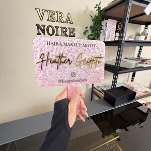 May include: A pink and gold business sign for "Vera Noire Hair & Makeup Artist Heather Griffith". The sign is held up by a person. The sign has a pink textured background and gold lettering. The Instagram handle @happyhairlady is also on the sign.