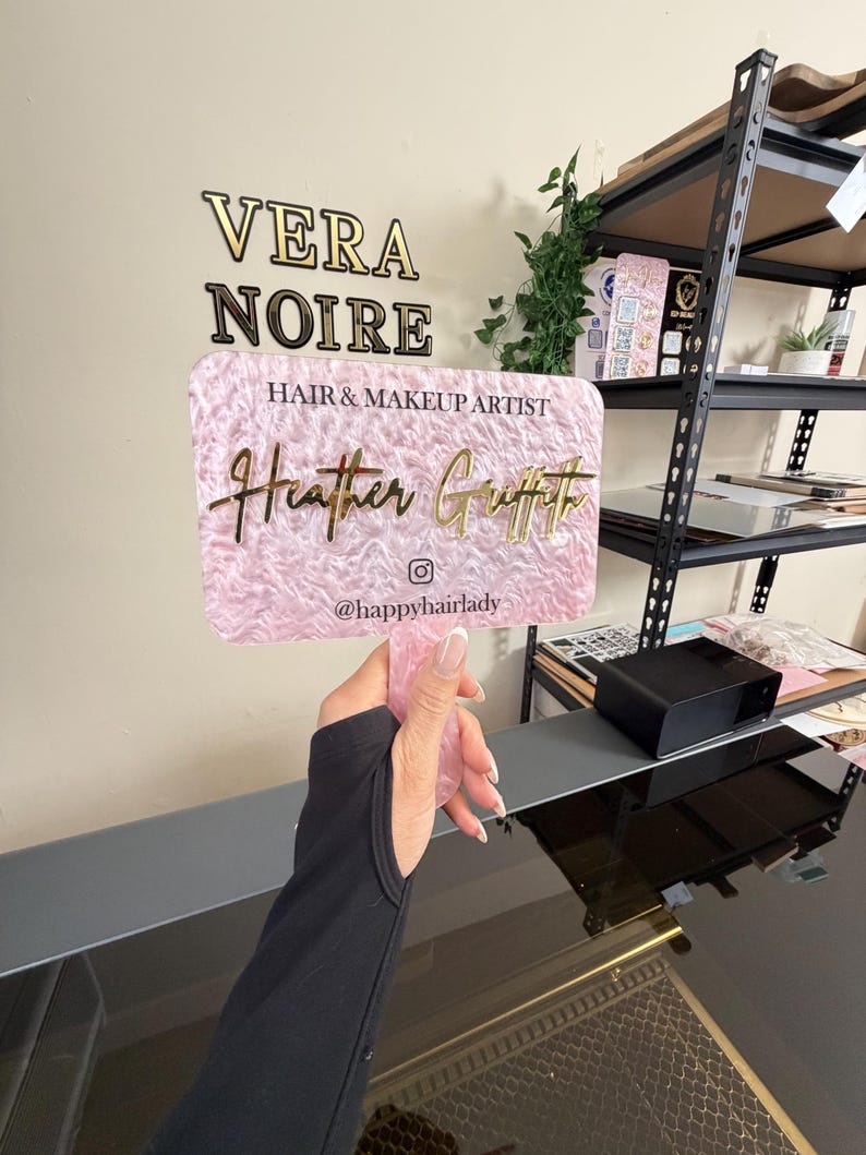 May include: A pink and gold business sign for "Vera Noire Hair & Makeup Artist Heather Griffith". The sign is held up by a person. The sign has a pink textured background and gold lettering. The Instagram handle @happyhairlady is also on the sign.