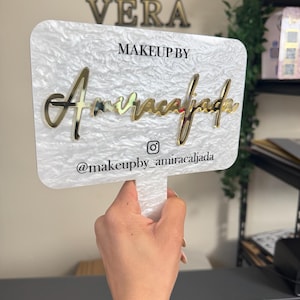 May include: A white and gold sign with the text "VERA" at the top. Below, it reads "MAKEUP BY" and "Amiracalfada" in gold script. The handle has the Instagram handle "@makeupby_amiracalfada".