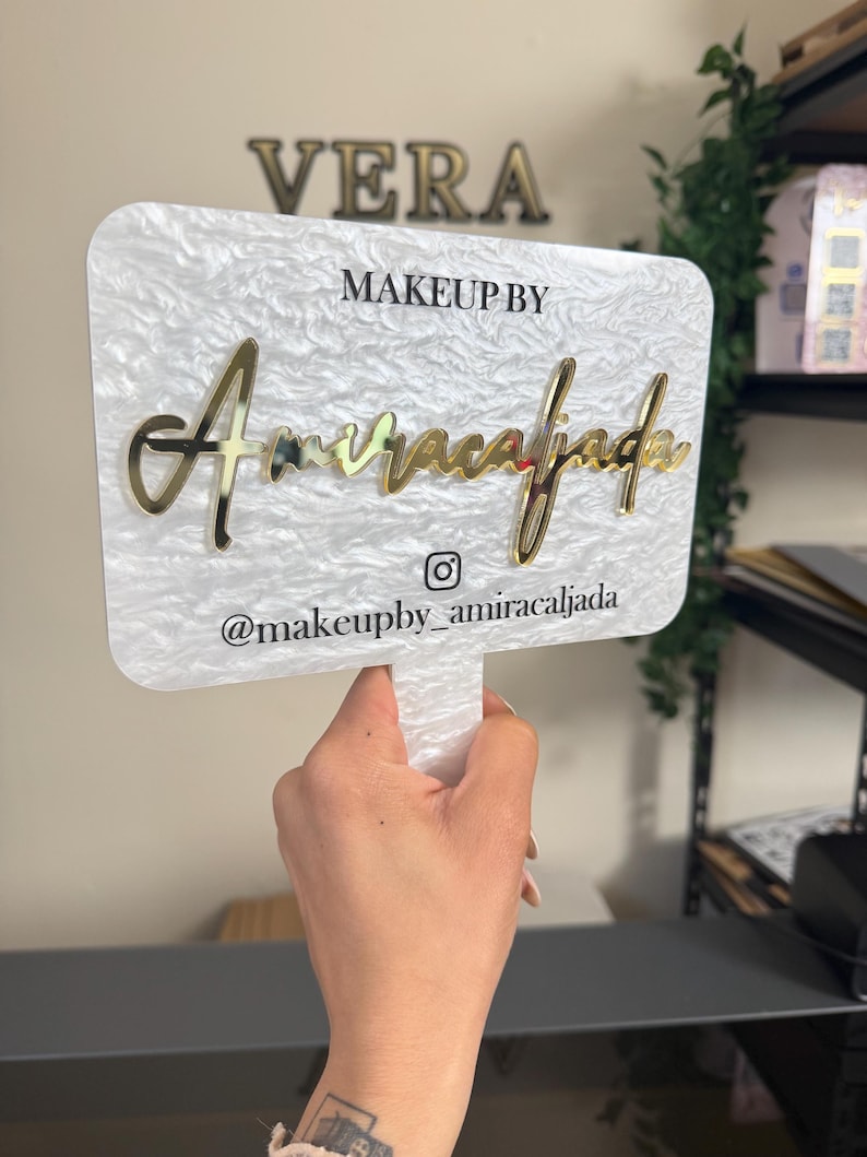 May include: A white and gold sign with the text "VERA" at the top. Below, it reads "MAKEUP BY" and "Amiracalfada" in gold script. The handle has the Instagram handle "@makeupby_amiracalfada".