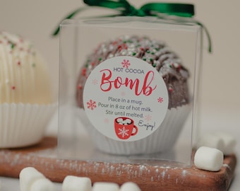 May include: A dark chocolate hot cocoa bomb in a clear square box with a green ribbon. The label reads: HOT COCOA Bomb, Place in a mug. Pour in 8 oz of hot milk. Stir until melted. Enjoy! A red mug graphic and snowflakes are also on the label.
