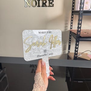 May include: A white and gold makeup mirror with the text "Makeup by Sarah Jaffer" and the Instagram handle "@saraidler_mua". The mirror has a marble-like pattern and is held up by a hand.