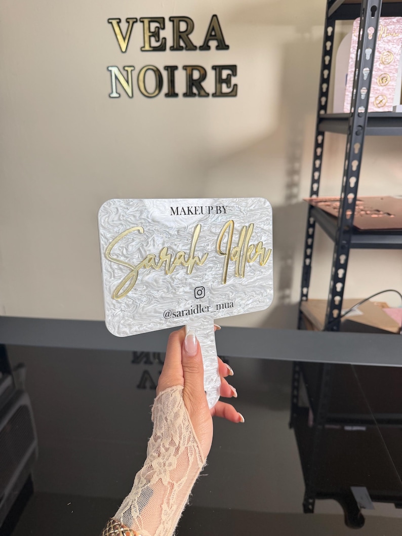 May include: A white and gold makeup mirror with the text "Makeup by Sarah Jaffer" and the Instagram handle "@saraidler_mua". The mirror has a marble-like pattern and is held up by a hand.