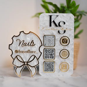 Puede incluir: Un letrero de negocios decorativo de acrílico en blanco y dorado. El letrero muestra la palabra "Nails" e iconos de redes sociales. El letrero está diseñado para mostrar información de contacto y perfiles de redes sociales.
