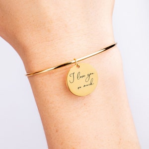 Op de afbeelding: Gouden armband met een ronde bedel waarop in cursief schrift "Ik hou van je zo veel" staat.