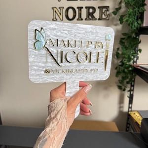 May include: A white and pearlescent sign with the text "MAKEUP BY NICOLE" in gold lettering, featuring a butterfly and makeup brush design. The sign also includes the Instagram handle @NICKIBEAUTY.CO.