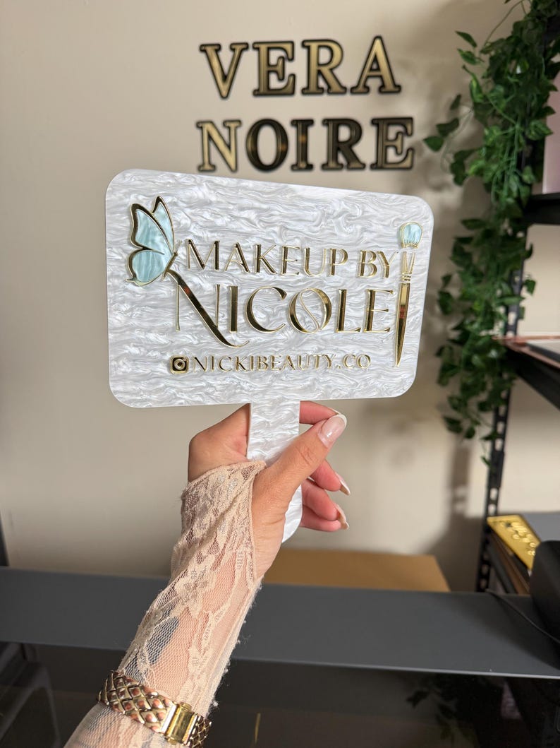 May include: A white and pearlescent sign with the text "MAKEUP BY NICOLE" in gold lettering, featuring a butterfly and makeup brush design. The sign also includes the Instagram handle @NICKIBEAUTY.CO.
