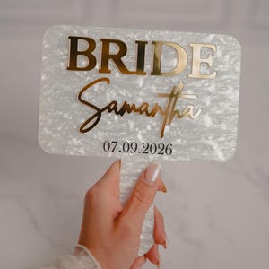 May include: A rectangular, pearlescent white sign with gold lettering that reads "BRIDE" and "Samantha" in a script font. The date "07.09.2026" is below. The sign is held by a hand.