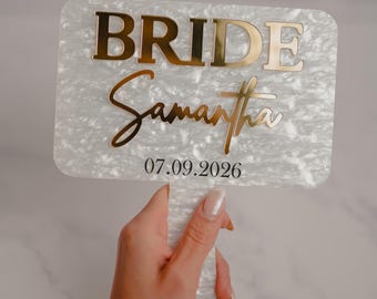 May include: A rectangular, pearlescent white sign with gold lettering that reads "BRIDE" and "Samantha" in a script font. The date "07.09.2026" is below. The sign is held by a hand.