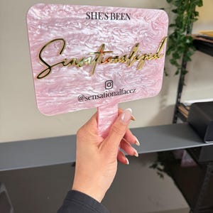 May include: A pink and white marbled acrylic sign with gold lettering that reads "She's Been Sensationalized" and the Instagram handle "@sensationalfacez". The sign is held by a hand.