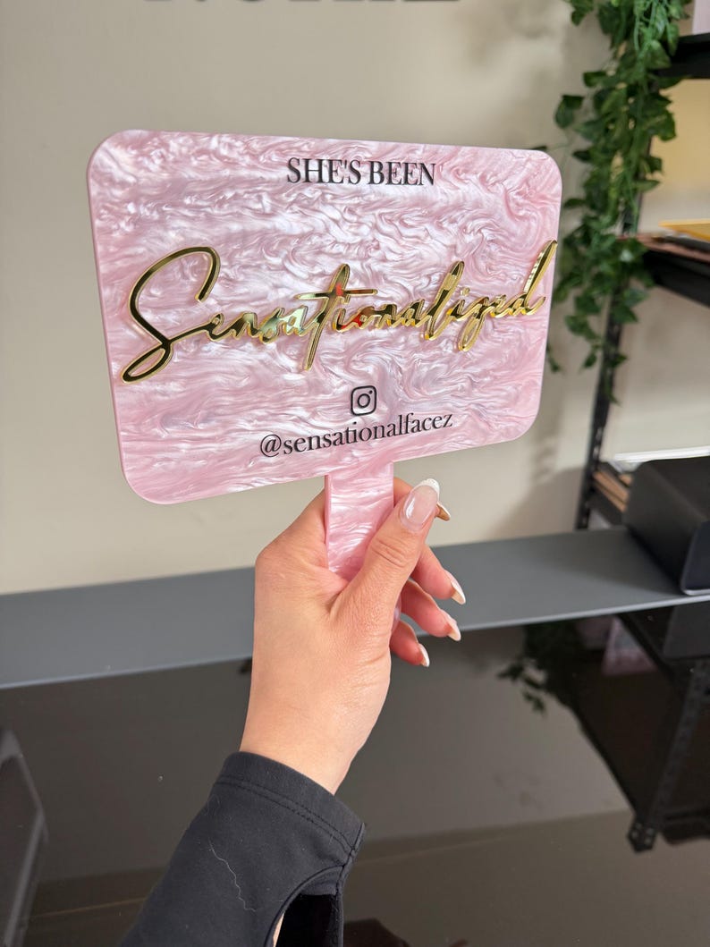 May include: A pink and white marbled acrylic sign with gold lettering that reads "She's Been Sensationalized" and the Instagram handle "@sensationalfacez". The sign is held by a hand.
