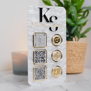 May include: A vertical white marbled acrylic sign featuring the black text "Ks" at the top. It includes three square QR codes with gold borders on the left and three circular gold social media icons on the right, all mounted on a matching base.