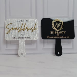 May include: Two makeup artist hand mirrors. One is white with the text "Makeup by Smashbrush" and the Instagram handle. The other is black with the text "ED BEAUTY" and the Instagram handle. Both have a handle.