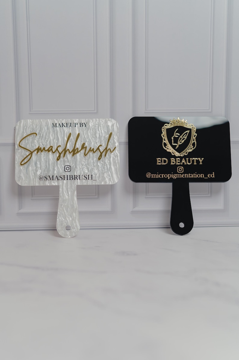 May include: Two makeup artist hand mirrors. One is white with the text "Makeup by Smashbrush" and the Instagram handle. The other is black with the text "ED BEAUTY" and the Instagram handle. Both have a handle.