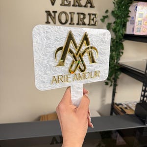 May include: A white, rectangular sign with a handle, featuring gold lettering that reads "ARIE AMOUR" and a gold emblem. The sign has a textured, marbled appearance. The words "VERA NOIRE" are displayed in gold letters on the wall behind the sign.